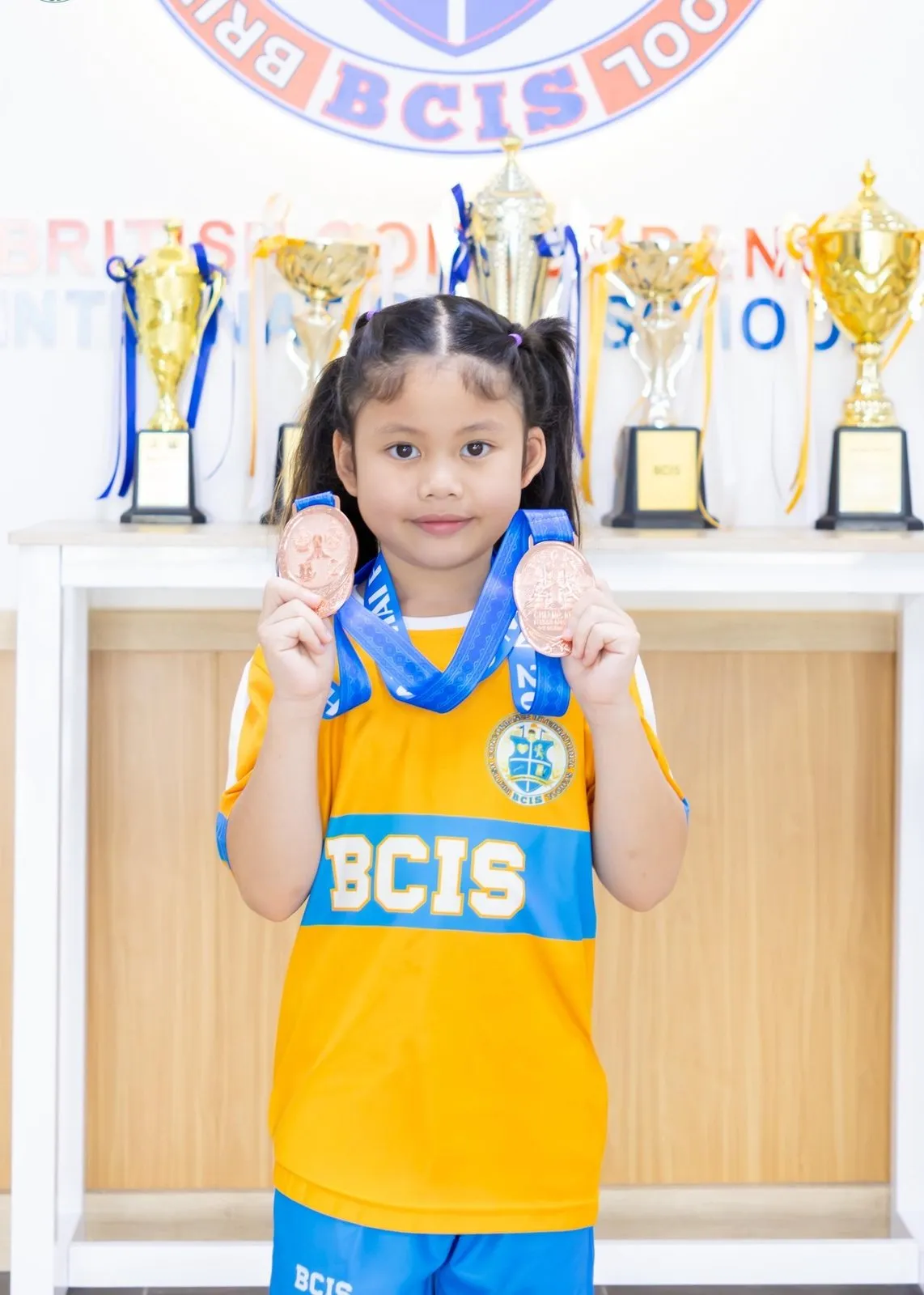 Primprim holding up her medals in front of the school trophies