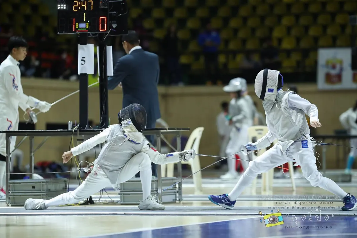 Two young fencers competing on the piste at the Nimibutr Sports Building, with an official and scoreboard in the background