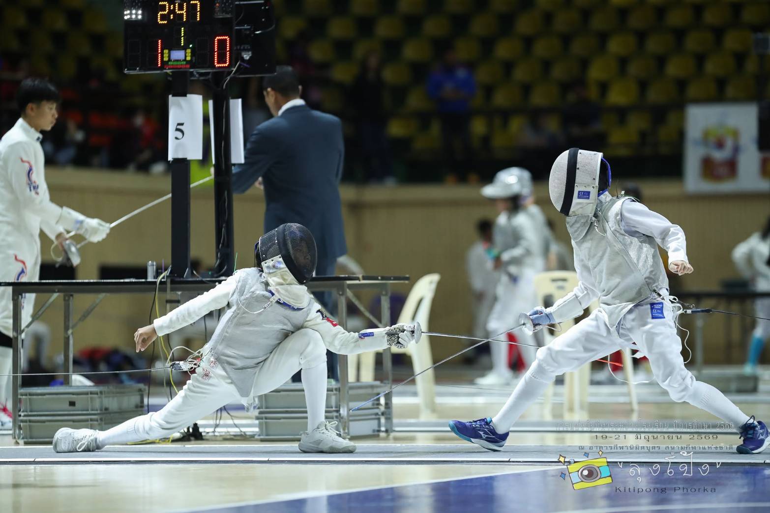 Two young fencers competing on the piste at the Nimibutr Sports Building, with an official and scoreboard in the background