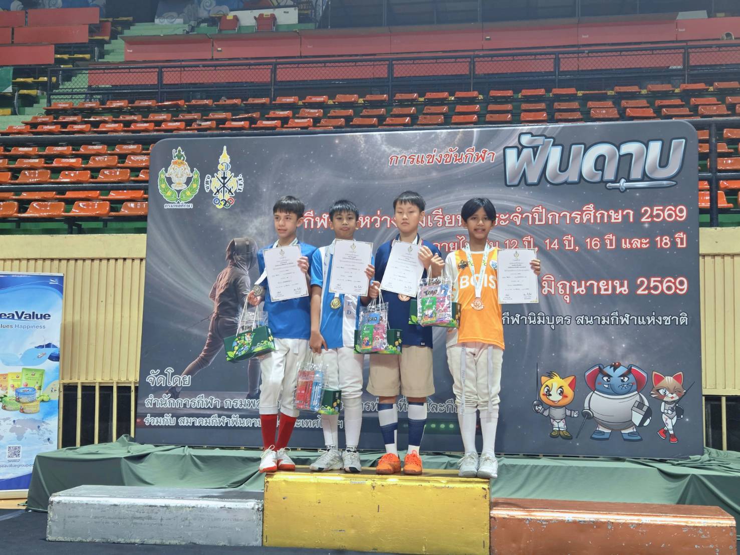 Pleum standing on the podium in his orange BCIS kit, holding up his bronze medal alongside three other young fencers