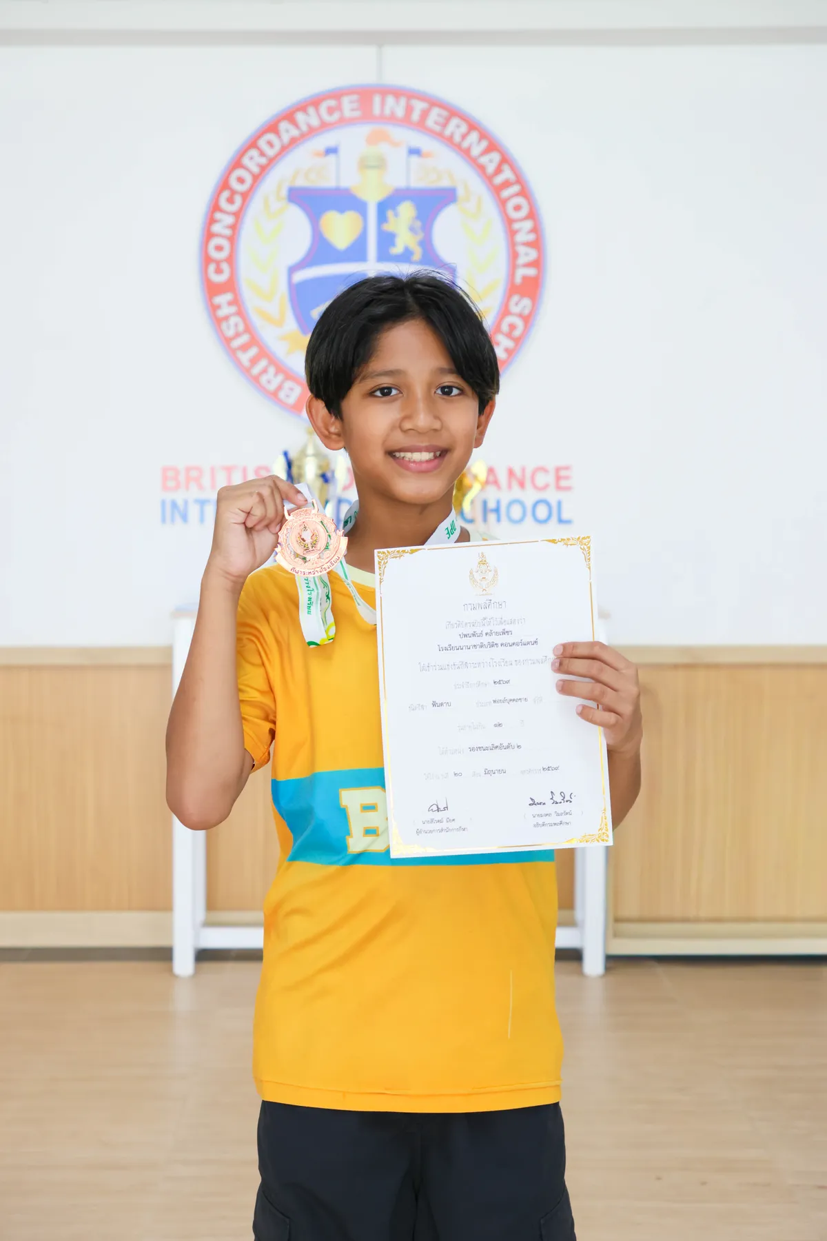 Pleum smiling and holding up his bronze medal and certificate in front of the BCIS crest at school