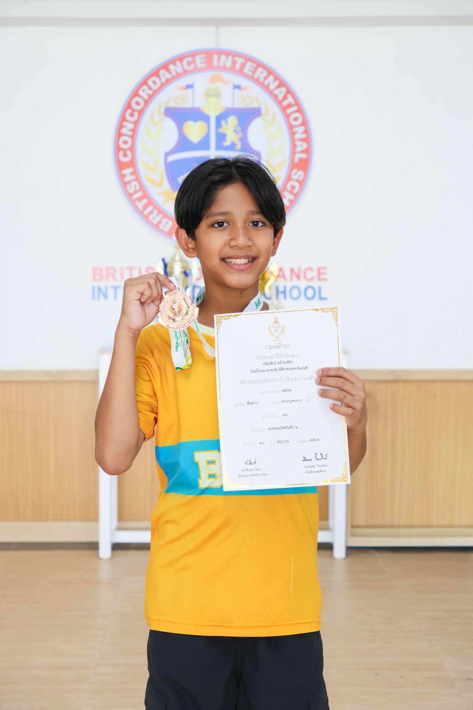 Pleum smiling and holding up his bronze medal and certificate in front of the BCIS crest at school