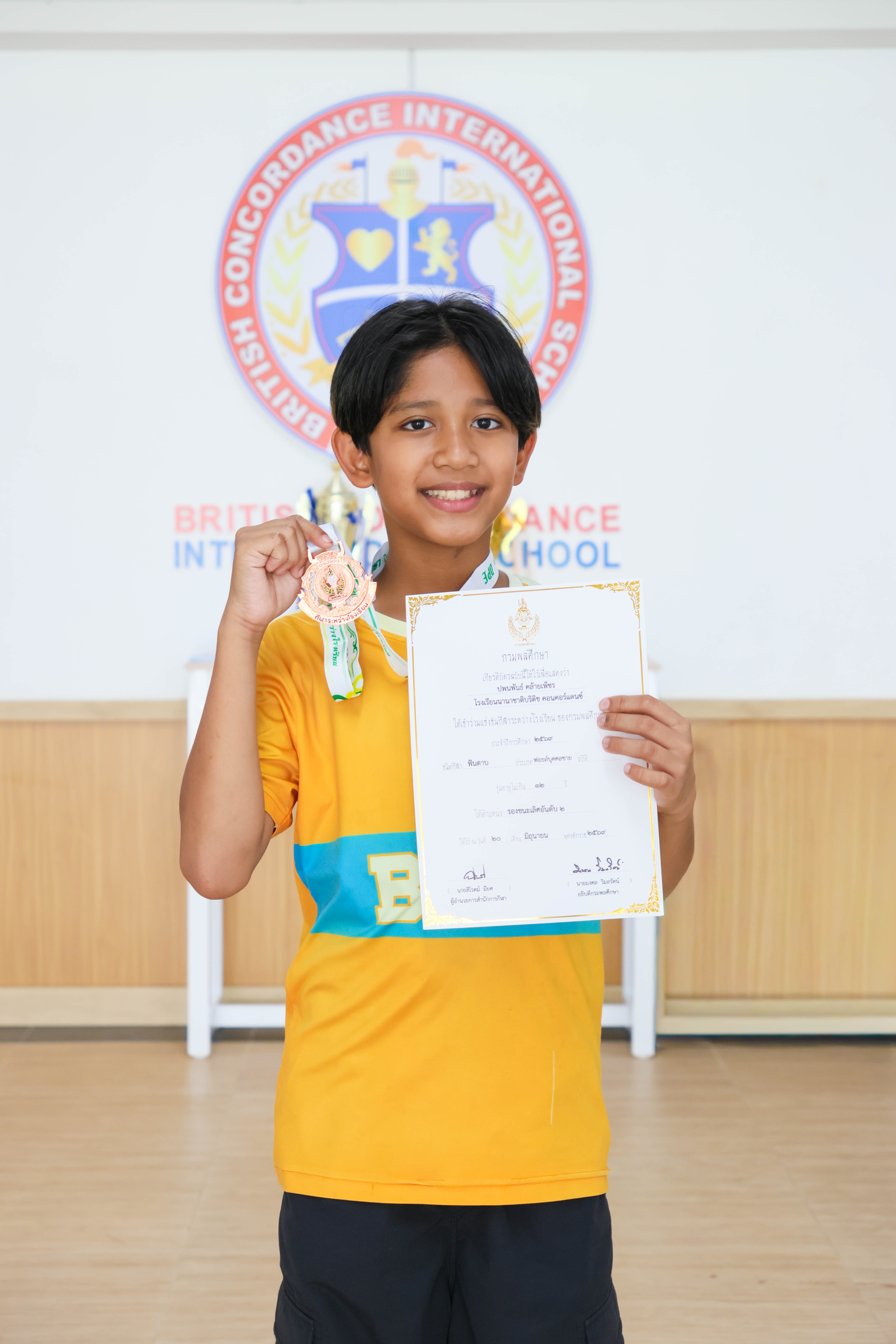 Pleum smiling and holding up his bronze medal and certificate in front of the BCIS crest at school
